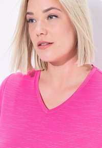Donna bionda con occhi azzurri che indossa un top fucsia a scollo a V, che guarda leggermente di lato su uno sfondo chiaro.