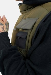 Chaleco acolchado con un exterior verde oliva y forro de polar negro. Presenta múltiples bolsillos: un bolsillo con solapa gris y un bolsillo negro.