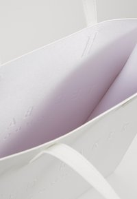 Sac fourre-tout blanc avec logo embossé, extérieur à texture lisse et doublure intérieure violette claire. Dispose de deux poignées robustes.