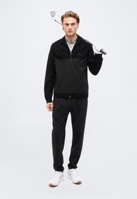 Jeune homme en veste noire zippée, polo gris, pantalon noir, gants blancs, tenant un club de golf sur l'épaule, debout sur fond blanc.