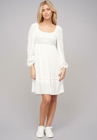 Vestido blanco con escote cuadrado, cuerpo texturizado, mangas largas abullonadas y falda acampanada. Combinado con zapatillas blancas.