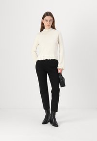 Un pull en tricot blanc avec un col montant et une finition en dentelle à l'ourlet, associé à un pantalon noir ajusté et des bottines noires élégantes.