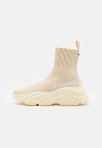 Baskets en style chaussette beige avec un dessus à côtes, une languette de tirage et une semelle en caoutchouc texturé. "Steve Madden" imprimé en blanc sur le côté.