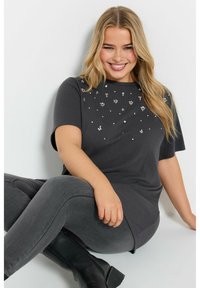 Grå kortärmad T-shirt med spridda strassdetaljer på framsidan, kombinerad med grå leggings och svarta ankelskor.