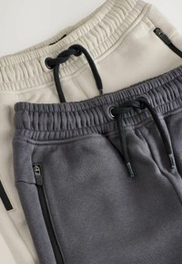 Two pairs of sweatpants: one beige and one dark gray, both with elastic waistbands, drawstrings, and zippered side pockets.