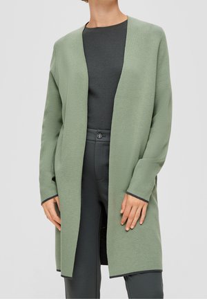 Personne portant un long cardigan vert clair ouvert à l'avant sur un haut gris foncé et un pantalon gris foncé avec une fermeture à bouton à la taille.