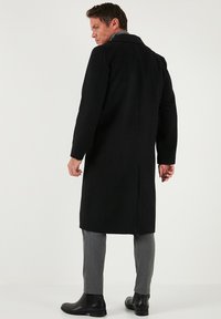Cappotto nero in lana con taglio dritto, spacco singolo e rever a lancia. Abbinato a pantaloni grigi e stivali eleganti neri. Design sartoriale e strutturato.
