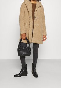 Beige kabát z umělého beránka s kapucí, nošený přes hnědý svetr, spárovaný s černými koženými kotníkovými botami a černou řasenou kabelkou.