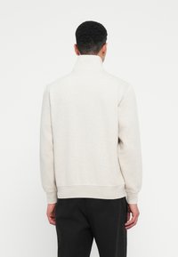 Άντρας φαίνεται από �πίσω φορώντας ένα ανοιχτό γκρι φούτερ με ψηλό λαιμό και μαύρο παντελόνι μπροστά σε λευκό φόντο.