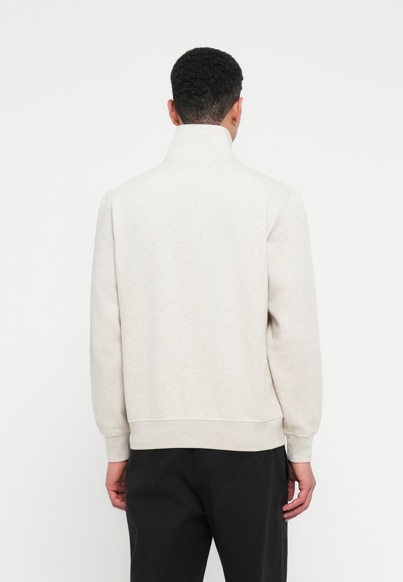 Ein Mann von hinten gesehen, der einen hellgrauen Pullover mit hohem Kragen und schwarze Hosen vor einem schlichten weißen Hintergrund trägt.