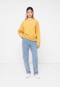 Gul genser med blomstermotiv, løs passform, ribbestrikkede mansjetter. Kombinert med lyseblå stripete jeans, avslappet snitt, og hvite sneakers.