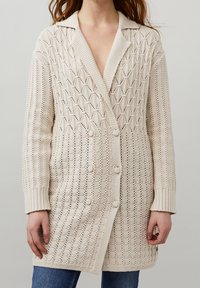 Gilet en tricot beige avec un motif en losanges, doté d'un col châle, de boutons double boutonnage et de poignets côtelés.