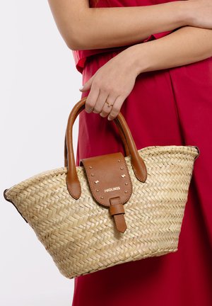 Mujer con vestido rojo sostiene un bolso de paja tejido con asas de cuero marrón y solapa etiquetada "Pikolinos" a la altura de la cintura.