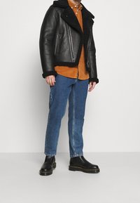 Giubbino da motociclista in pelle nera con colletto in shearling, abbinato a una camicia in corduroy arancione e jeans blu. Stivali neri fino alla caviglia completano l'outfit.