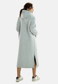 Sudadera larga gris hecha de una tela suave, con capucha, aberturas laterales y mangas sueltas, combinada con zapatillas blancas.