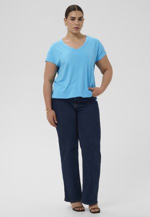 Vrouw staand met één hand in de zak, gekleed in een lichtblauw T-shirt met V-hals, donkerblauwe spijkerbroek, zwarte sandalen met hak en een armband om de linkerpols.