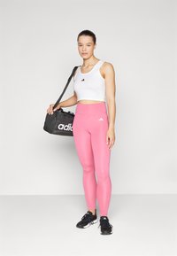 Top desportivo branco cropped com logo preto, leggings cor-de-rosa e sapatilhas pretas. O modelo segura uma mala de ginásio preta com um logo branco.