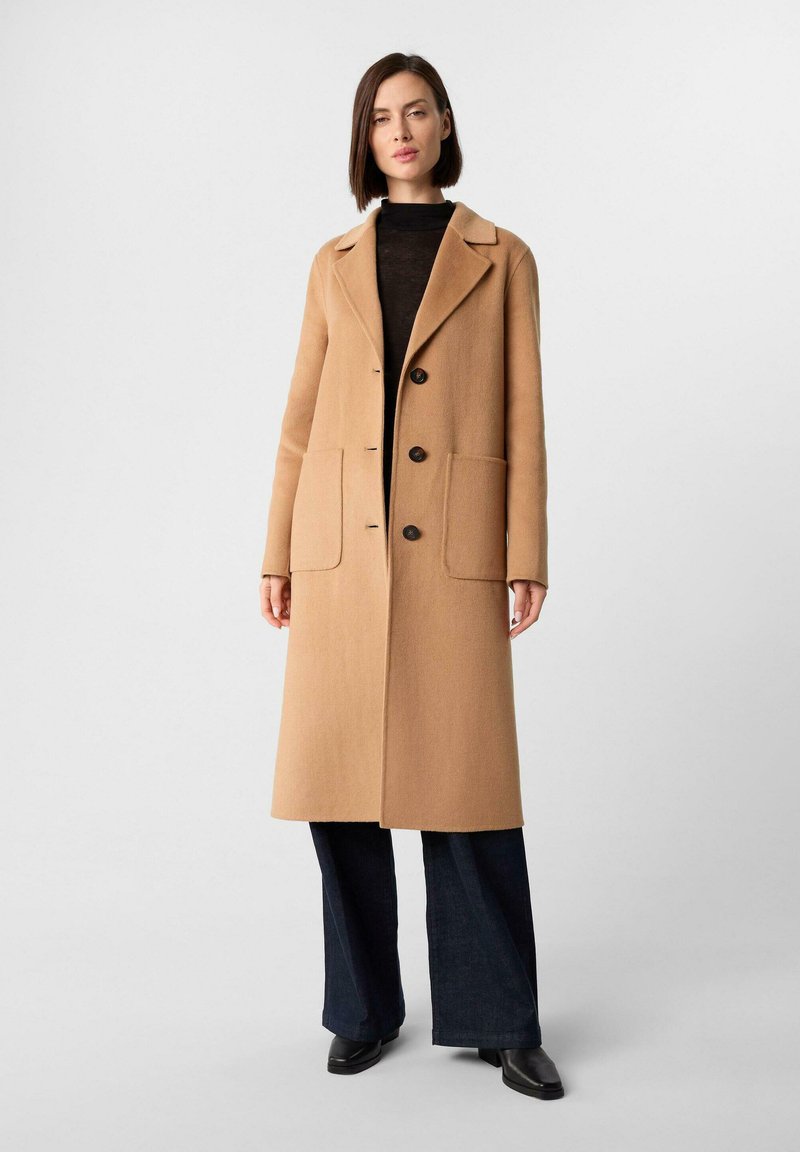 Donna che indossa un lungo cappotto color cammello con ampie tasche frontali, dolcevita nero, pantaloni larghi scuri e scarpe nere, in piedi contro uno sfondo semplice.