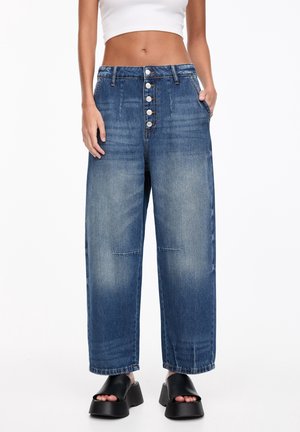 Vrouw draagt een blauwe denim spijkerbroek met wijde pijpen en hoge taille, met knoopsluiting aan de voorkant, en zwarte platform sandalen met open teens, staand tegen een witte achtergrond.