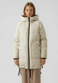 Beige gewatteerde parka met een ritssluiting aan de voorkant, grote capuchon, zijzakken en zwarte hardware accenten. Voorzien van trekkoorden aan de onderkant voor verstelbaarheid.