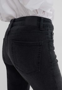 Jean en denim noir avec une taille haute, des poches arrière et une finition matte texturée. Présente une étiquette logo près de la taille.