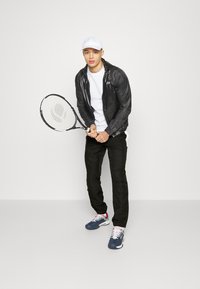 Svart lett jakke med teksturert overflate, hvit t-skjorte, svarte bukser, marineblå sko, holder en tennisracket, hvit caps.
