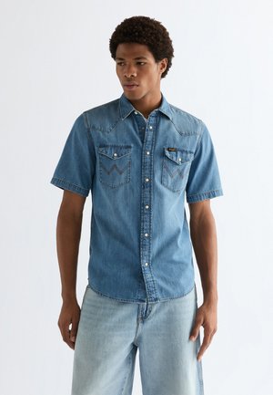Homme portant une chemise en denim bleue à manches courtes avec des poches poitrine boutonnées et un jean bleu clair, debout devant un fond clair uni.
