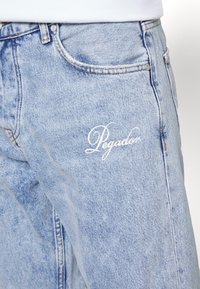 Ljusblå denimjeans med en urtvättad textur, med vit broderad text "Pegador" på det främre fickområdet. Klassisk femficksdesign.