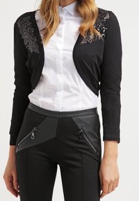 Donna che indossa un cardigan nero corto con dettagli in paillettes sopra una camicia bianca con bottoni e pantaloni neri con tasche con cerniera.