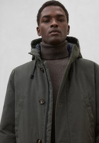 Ciemnozielona kurtka z kapturem, zapinana na guziki i z sznurkiem do regulacji, noszona na brązowym, prążkowanym golfie. Charakteryzuje się gładką teksturą i praktycznym designem.