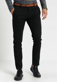 Man som bär svarta smala byxor, brunt bälte, svarta sneakers och en blå mönstrad långärmad skjorta med ena handen i fickan.