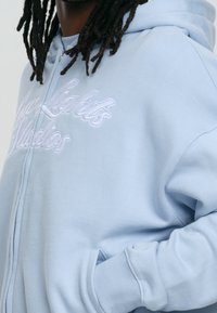 Sudadera con cremallera de color azul claro, hecha de tela suave, con texto bordado en blanco y bolsillos tipo canguro. Textura suave con puños acanalados.