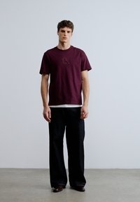 Burgundy cotton t-shirt with a raised logo on the chest, paired with black wide-leg trousers and brown shoes. Simple, relaxed fit.