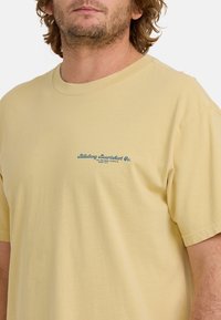 Jasnożółta bawełniana koszulka z okrągłym dekoltem, krótkimi rękawami i niebieskim nadrukiem logo po lewej stronie klatki piersiowej. Miękka, gładka struktura.