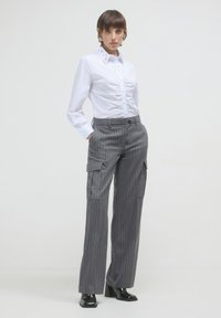 Donna che indossa una blusa bianca arricciata con collare impreziosito, pantaloni cargo grigi a righe e stivaletti neri con tacco a blocco, in piedi con le mani in tasca.
