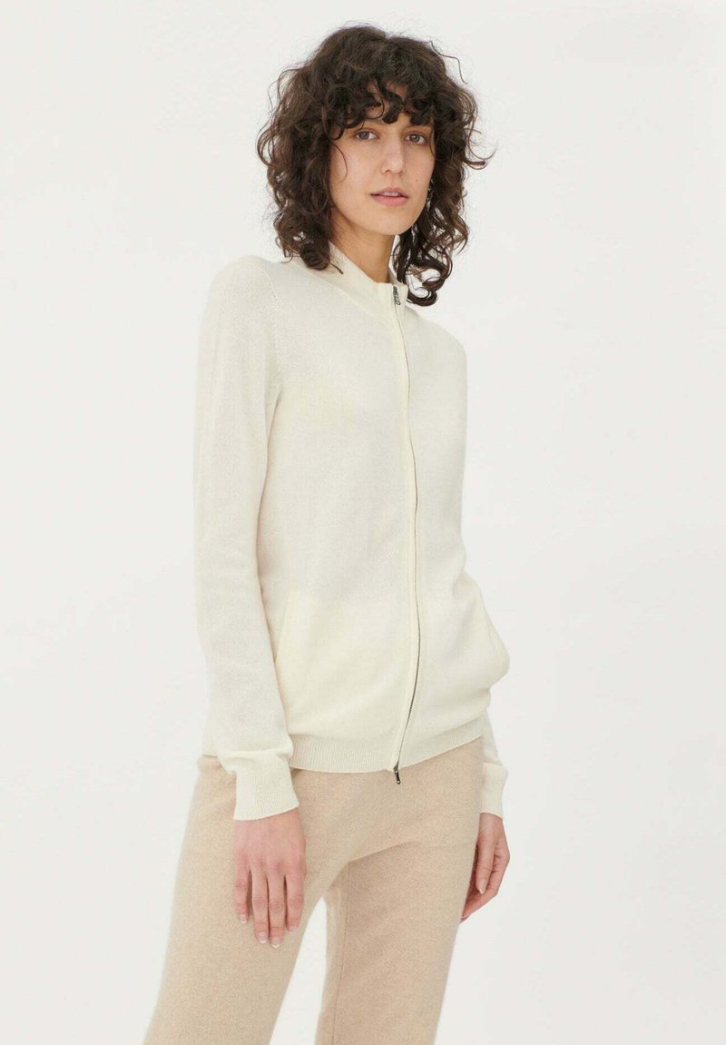 Långärmad krämfärgad zip-up cardigan i mjukt tyg, med hög krage, framfickor och ribbade muddare och fåll.