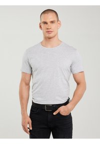 Mand med kort hår iført en lysegrå tætsiddende t-shirt, sort bælte og sorte jeans, stående mod en ensfarvet lys baggrund.