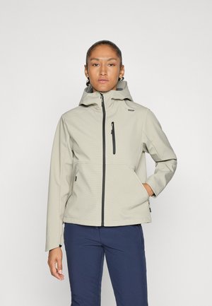 Person standing with hand in pocket wearing light gray hooded jacket and dark blue pants against plain light background.