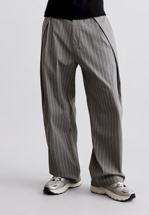 Grijze broek met brede pijpen en fijne strepen, gecombineerd met zilvergrijze sportschoenen bij een staand persoon tegen een effen achtergrond.