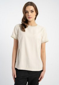 Beżowa bluzka z krótkim rękawem i okrągłym dekoltem, zdobiona misterną koronką na rękawach i dekolcie. Gładki materiał o luźnym kroju.