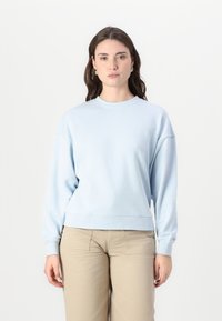 Felpa azzurra chiara realizzata in tessuto morbido, con colletto rotondo, spalle scese e polsini a costine, abbinata a pantaloni beige.