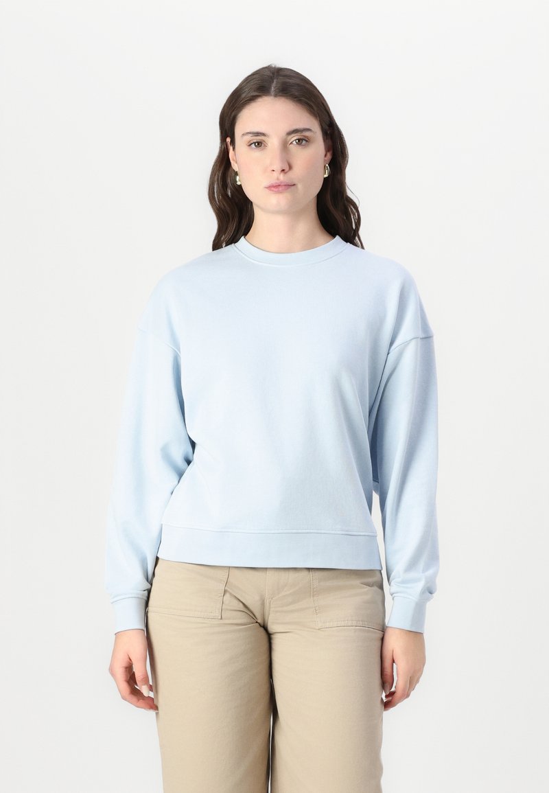 Felpa azzurra chiara realizzata in tessuto morbido, con colletto rotondo, spalle scese e polsini a costine, abbinata a pantaloni beige.