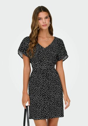 Nő fekete, fehér pöttyös rövid ujjú ruhában, fekete kézitáskát tart, hullámos barna haja van, és minimális ékszereket visel.