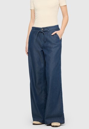 Model trägt weit geschnittene dunkelblaue Jeans mit Kordelzug und Knopf am Bund, kombiniert mit beigen Schuhen und einem hellbeigen kurzärmeligen Oberteil.