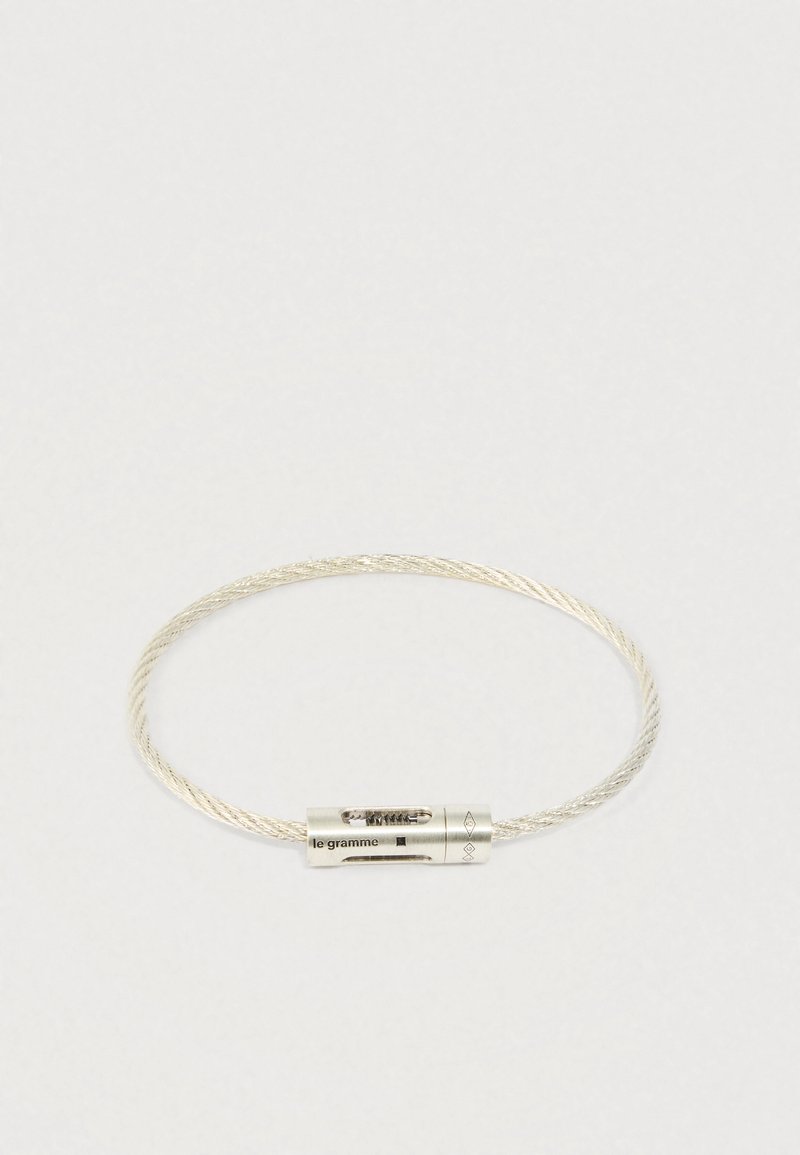 Sudraba vijīga vadu aproce ar cilindrisku aizdari, uz kuras ir iegravēts "le gramme" uz baltas fona.