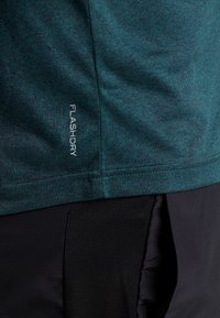 Camiseta deportiva en color azul verdoso con un tejido texturizado y un patrón sutil. Logo "FLASHDRY" impreso verticalmente en blanco en el lado izquierdo.