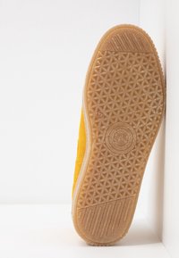 Semelle d'une chaussure jaune avec une semelle extérieure en caoutchouc texturé présentant des motifs géométriques et des lignes d'adhérence, photographiée sur un fond blanc.