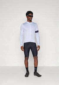 Vyras vilkintis šviesiai mėlyną ilgomis rankovėmis dviratininko marškinį, juodus dviratininko šortus, juodas kojines, juodus dviratininko batelius ir juodus akinius nuo saulės.