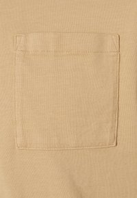 Beige tričko z látky s texturovaným povrchem, které má našitou kapsu na levé straně. Měkký materiál a jednoduché designové detaily.