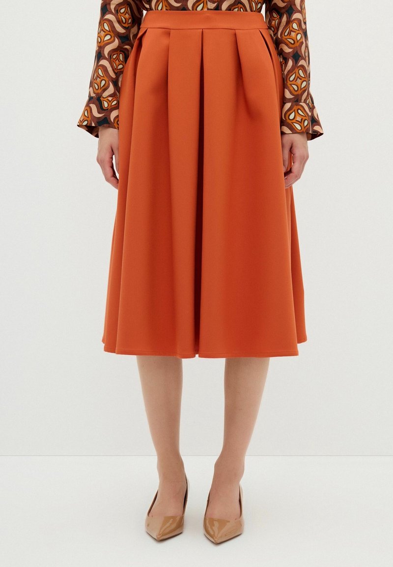 Rust-orange midi kjol med veck, gjord av slätt tyg. Har hög midja och når under knäna, kombinerad med beige spetsiga klackar.